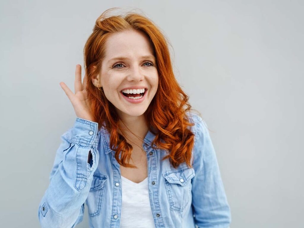 Lächelnde Frau mit roten Haaren in Jeanshemd, hält Hand am Ohr. Freundlichkeit und positive Ausstrahlung im Porträt.