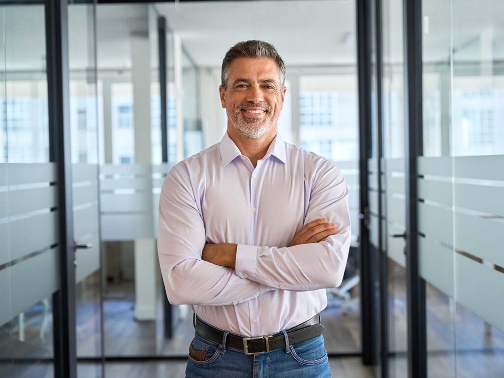 Mann im Büro mit verschränkten Armen, lächelnd, lässiges Hemd. Moderne Arbeitsumgebung, Führungskraft, Business-Kontext.