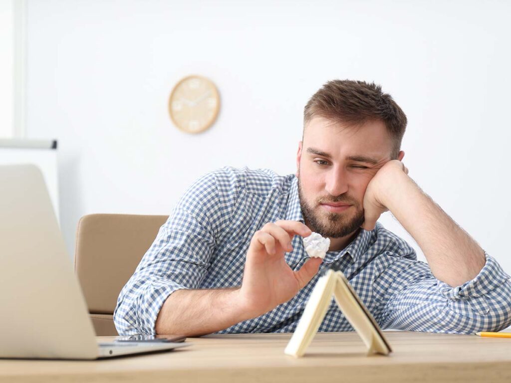Mann in kariertem Hemd am Schreibtisch, betrachtet nachdenklich Papierball, Laptop geöffnet. Kreative Blockade, Büroarbeit.