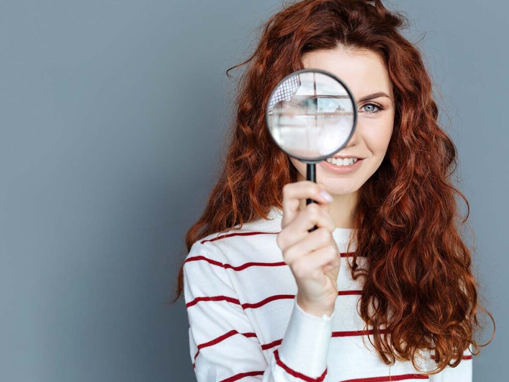 Rothaarige Frau in gestreiftem Pullover blickt durch eine Lupe. Konzept für Neugier, Suche und Entdeckung.