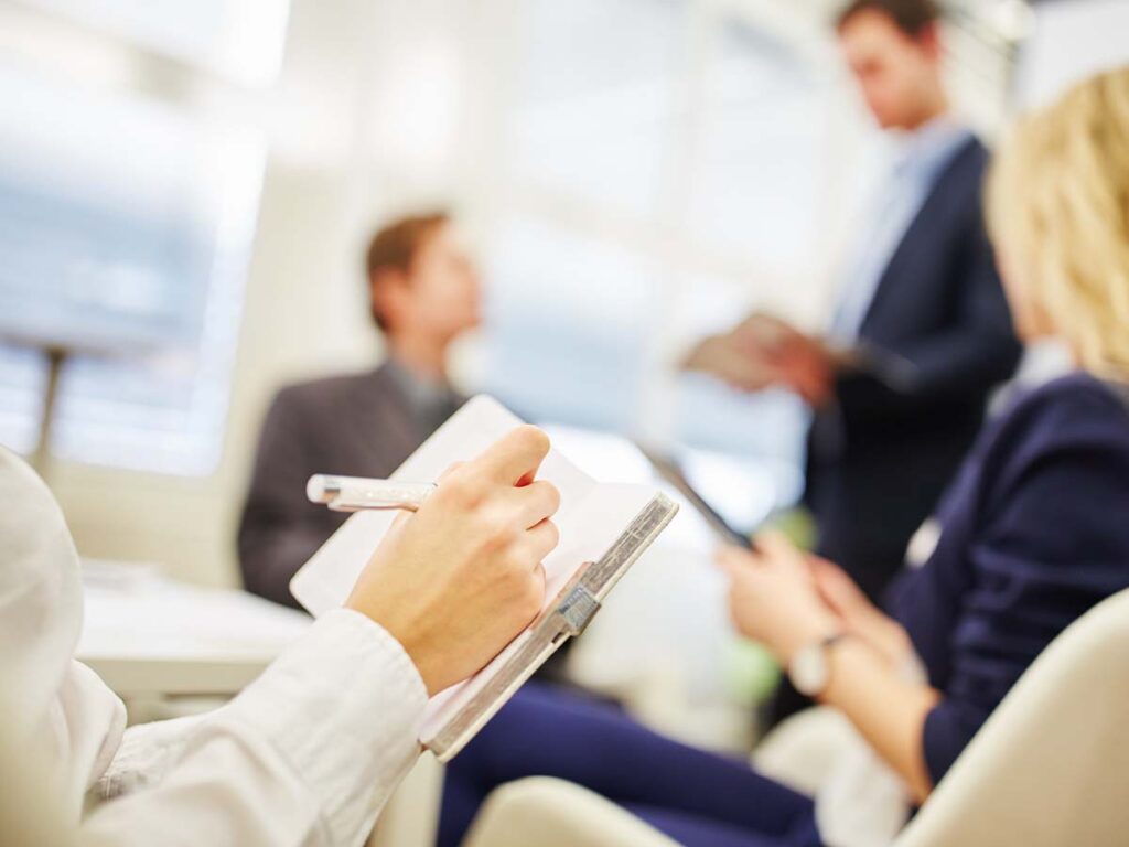 Notizen während eines Meetings im Büro, Personen im Hintergrund, Schwerpunkt Teamarbeit und Geschäftsstrategie.