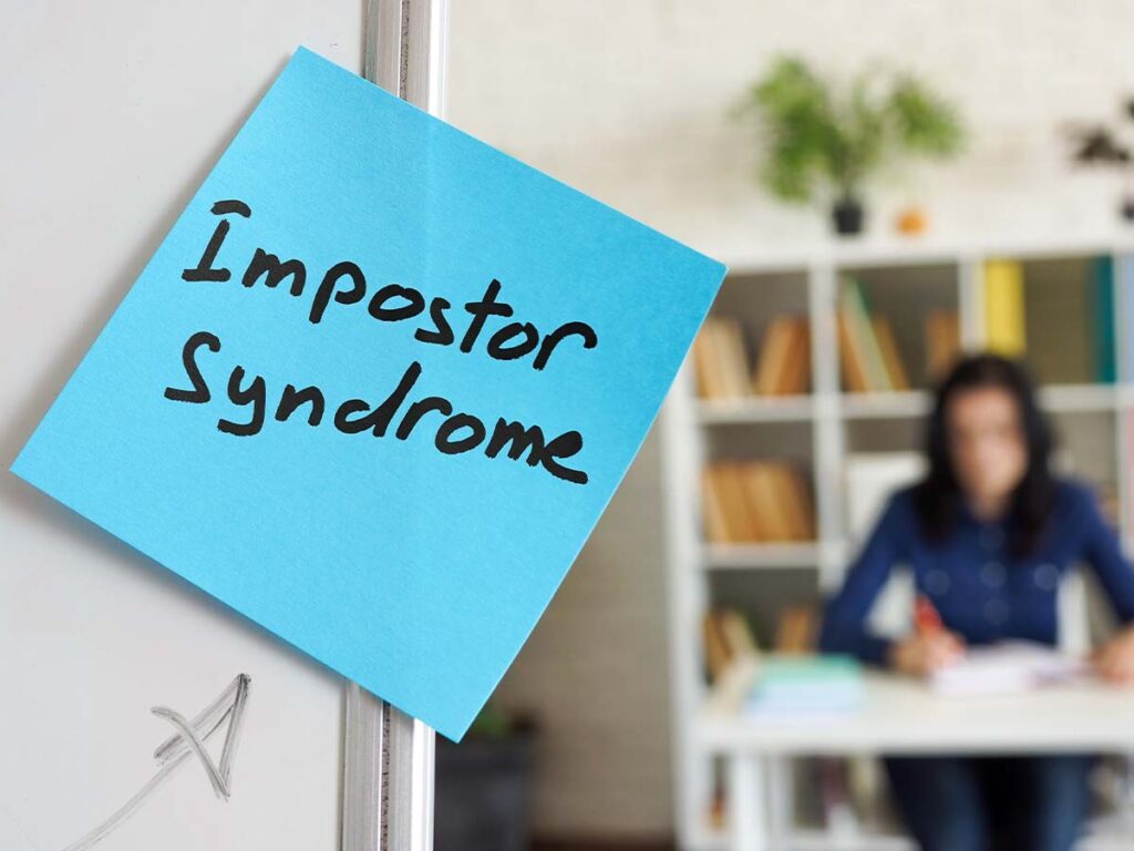 Blauer Zettel mit Impostor Syndrome, unscharfer Hintergrund Frau am Schreibtisch, Konzept für Selbstzweifel und Psychologie.