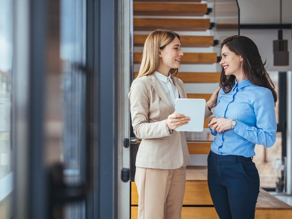 Zwei Geschäftsfrauen unterhalten sich freundlich im modernen Büro mit Tablet. Karriere und Networking.