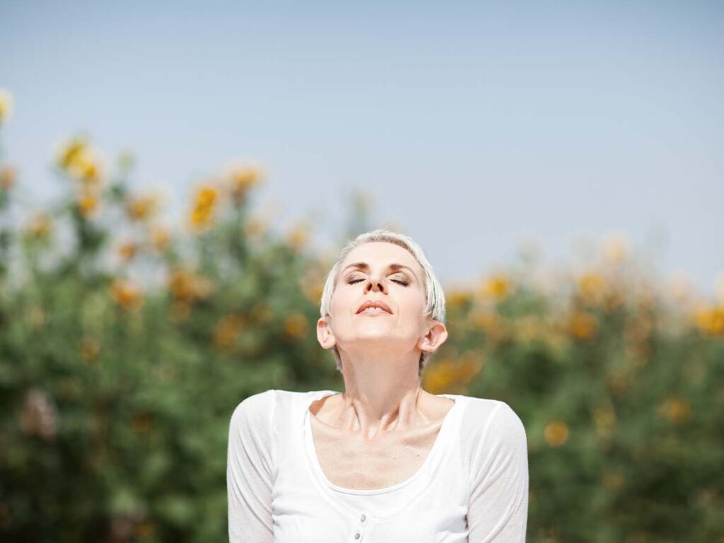 Frau genießt sonniges Wetter in einem Feld voller blühender Sonnenblumen, mit geschlossenen Augen und entspanntem Gesichtsausdruck.