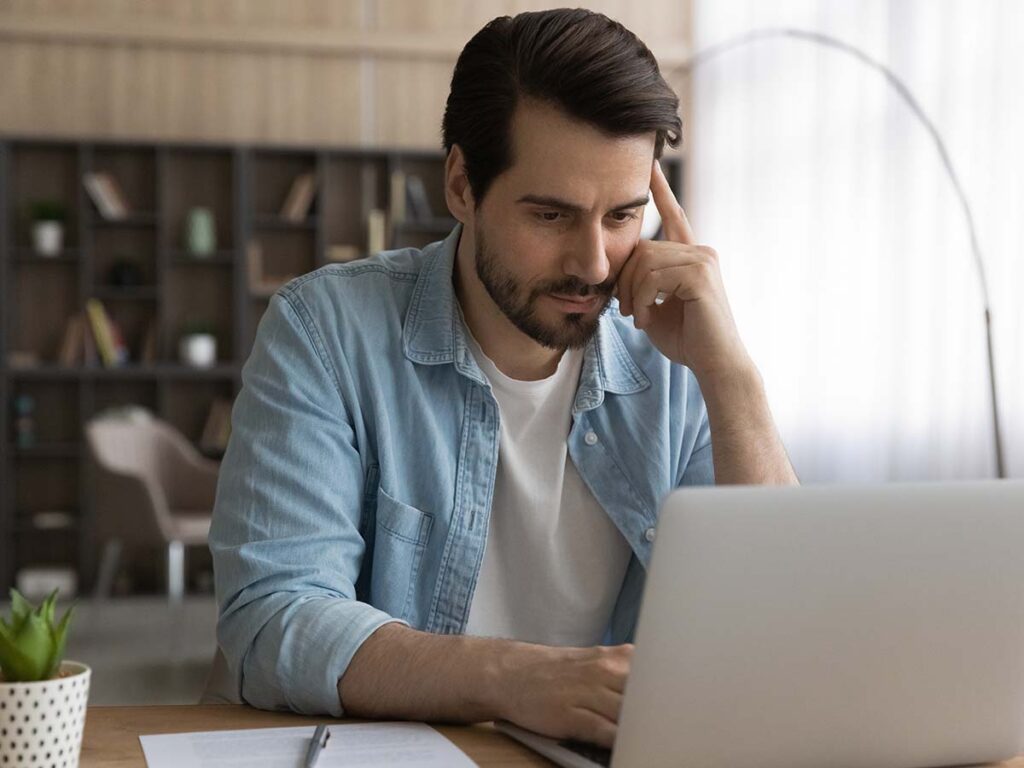 Mann konzentriert am Laptop, Homeoffice-Umgebung, Bücherregal im Hintergrund, casual Kleidung, Tageslicht, Arbeiten von zu Hause.