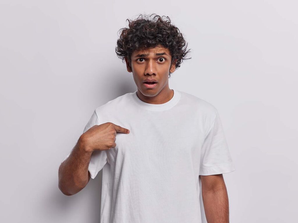 Ein überraschter Mann mit lockigem Haar trägt ein weißes T-Shirt und zeigt auf sich selbst, vor neutralem Hintergrund.