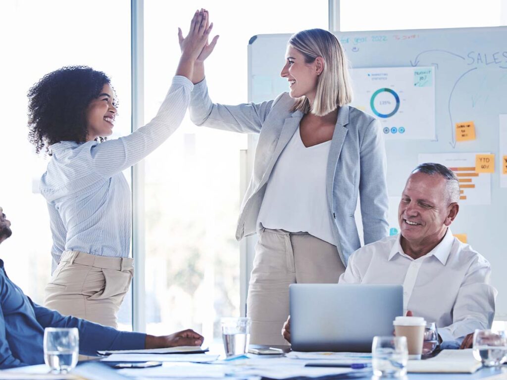 Team-Meeting: Zwei Geschäftsfrauen geben ein High-Five, während ein Kollege am Laptop arbeitet. Zusammenarbeit und Erfolg.