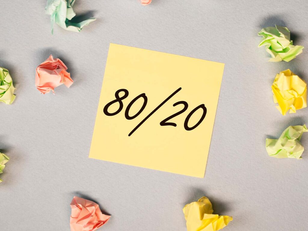 Gelber Zettel mit der Aufschrift 80/20, umgeben von bunten, zerknüllten Papierbällen; Konzept des Pareto-Prinzips.