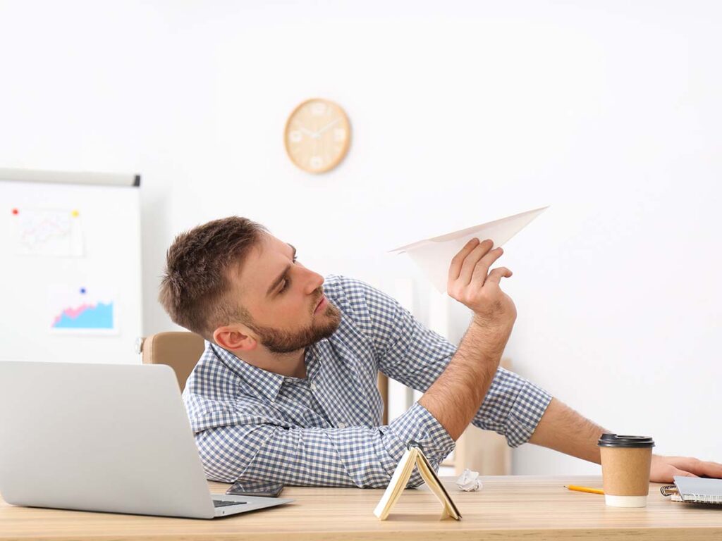 Mann im Büro wirft Papierflieger, Laptop und Kaffeebecher auf Schreibtisch. Kreativität und Pause bei der Arbeit.