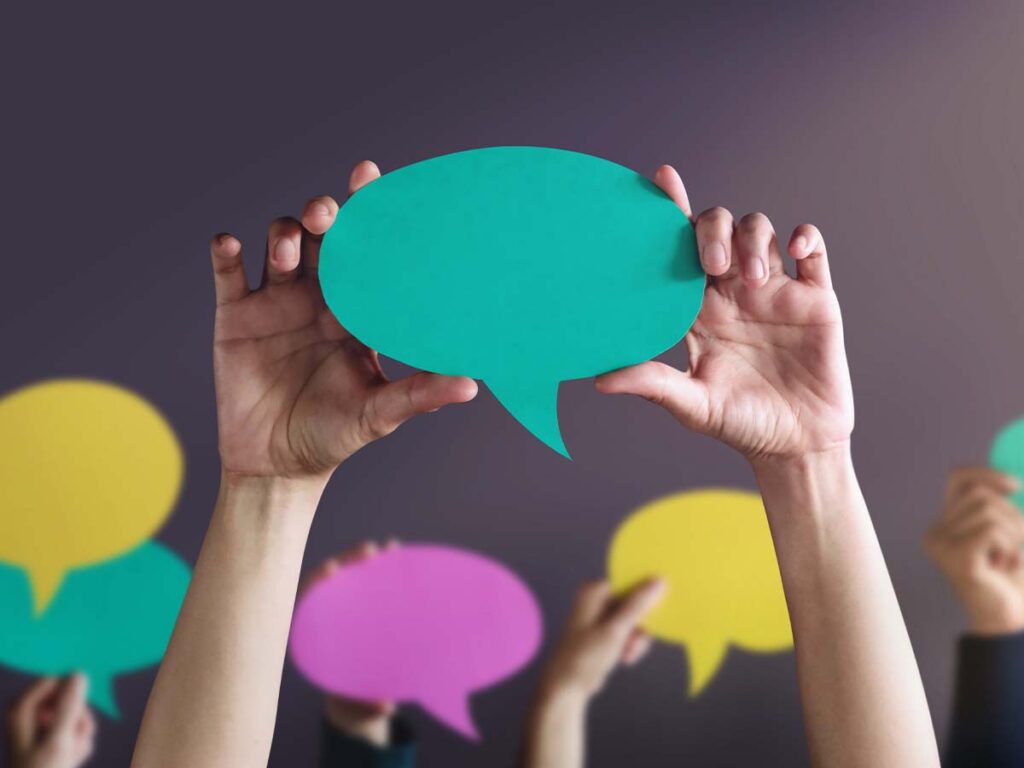 Hände halten bunte Sprechblasen in die Luft, symbolisierend Kommunikation und Zusammenarbeit bei einem Meeting oder Workshop.