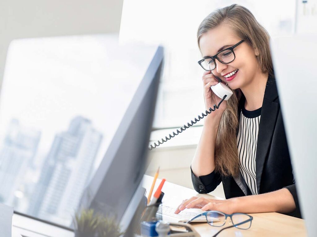 Frau im Büro telefoniert lächelnd am Schreibtisch, nutzt Computer. Moderne Geschäftskommunikation, Büroarbeit, Callcenter.