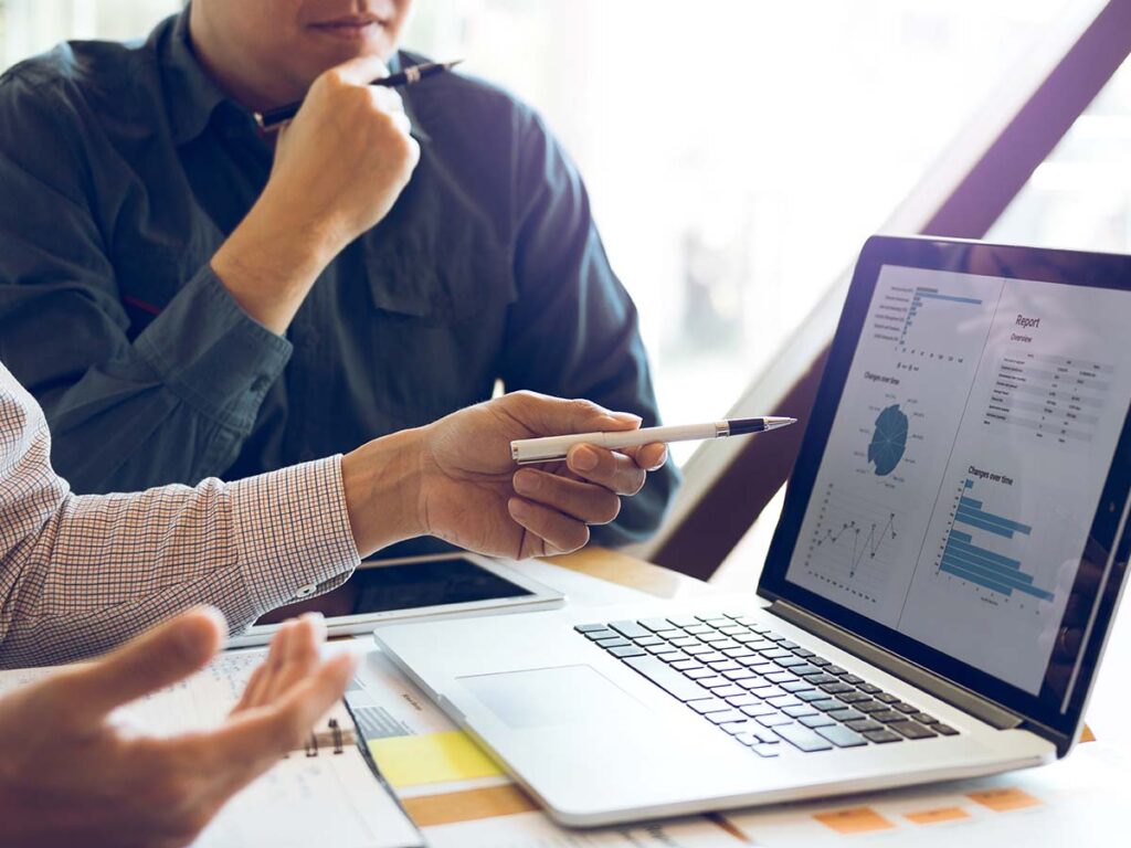 Personen diskutieren Geschäftsdaten auf einem Laptop-Bildschirm. Grafiken und Tabellen zeigen Analyse- und Marktforschungsergebnisse.