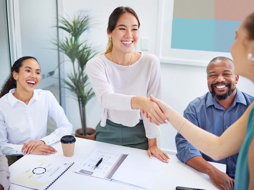 Frau in Büro schüttelt Händeschlag mit Kollegin, während Team lächelnd zusieht. Teamwork und Zusammenarbeit im Fokus.
