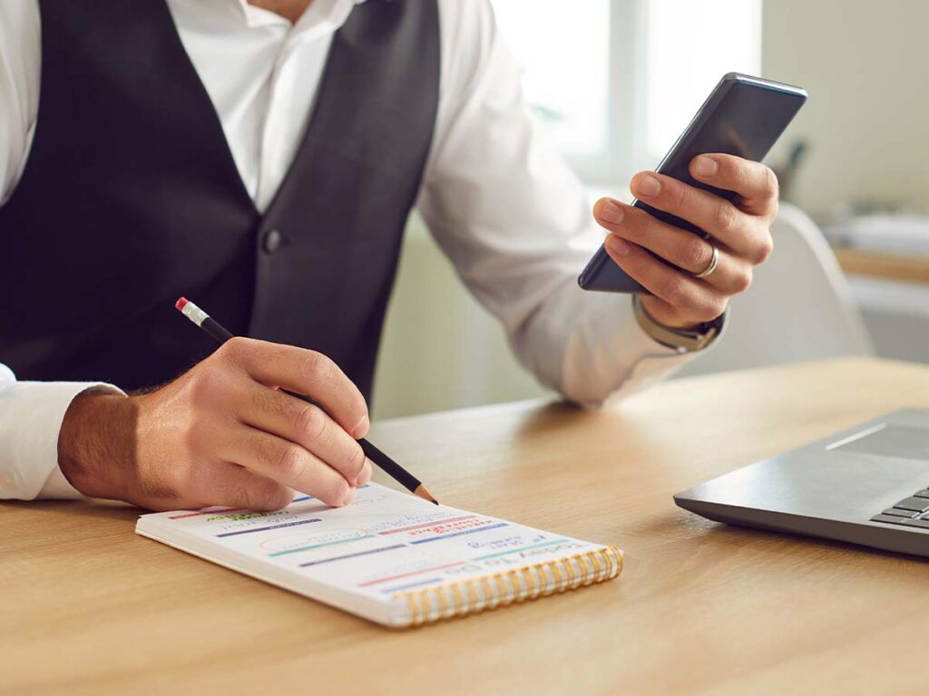 Mann in Business-Kleidung mit Smartphone überprüft Notizen am Schreibtisch; produktives Arbeiten und Multitasking im Büro.