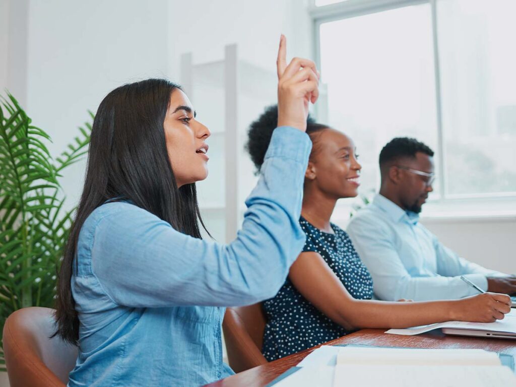 Person hebt Hand im Meeting, um Frage zu stellen. Multikulturelle Gruppe, Teamarbeit, Diskussion, Büro, Zusammenarbeit.