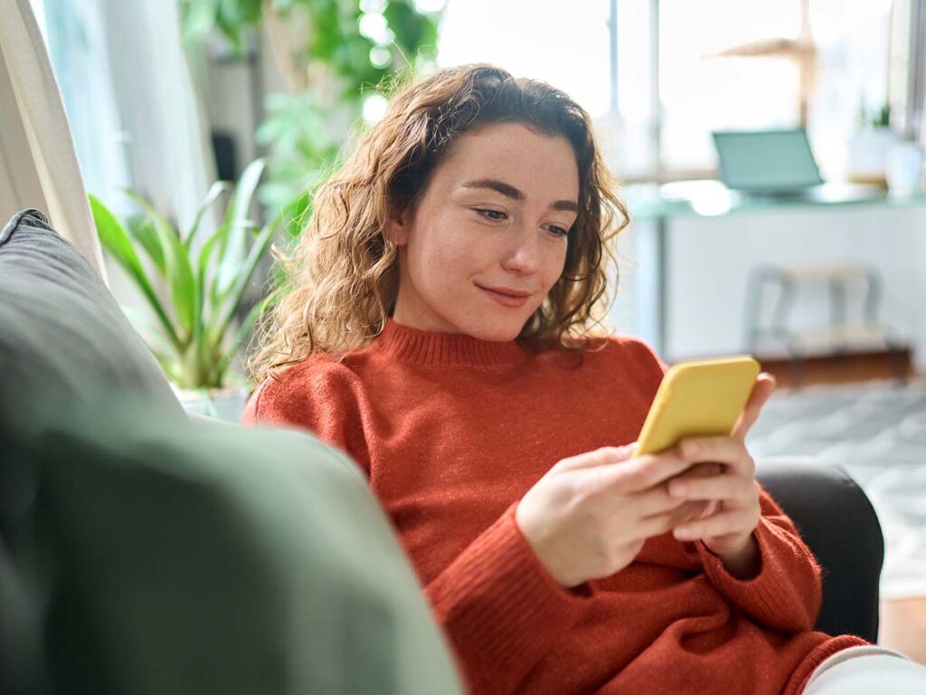 Frau in rotem Pulli sitzt auf dem Sofa und schaut entspannt aufs Smartphone. Innenraum mit Pflanzen im Hintergrund.
