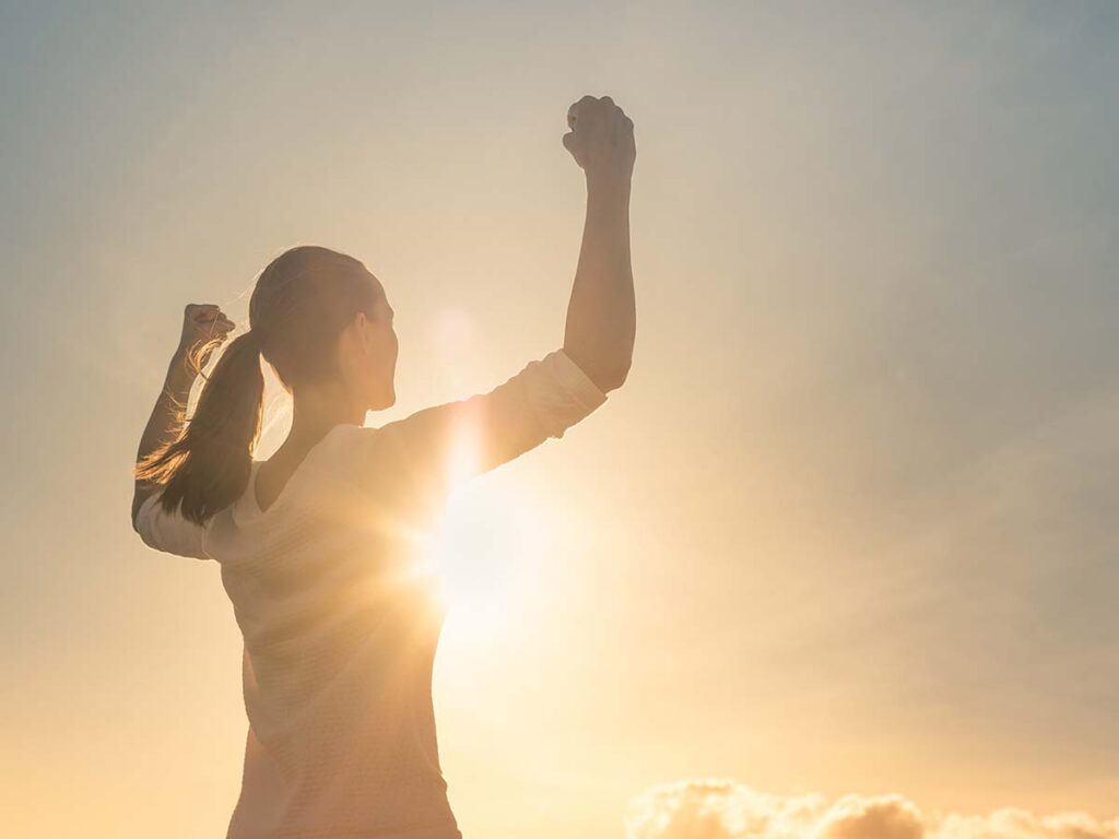 Frau bei Sonnenaufgang, hebt Arme siegreich. Symbol für Erfolg, Motivation und Freiheit. Ideal für Lebensstil und Fitness-Themen.