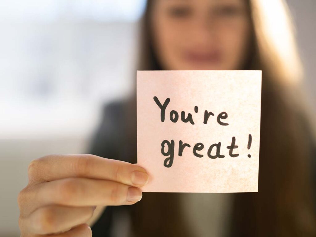 Frau hält Notiz mit You're great! in der Hand. Motivation und Zuspruch durch handgeschriebenes Lob auf Papier.