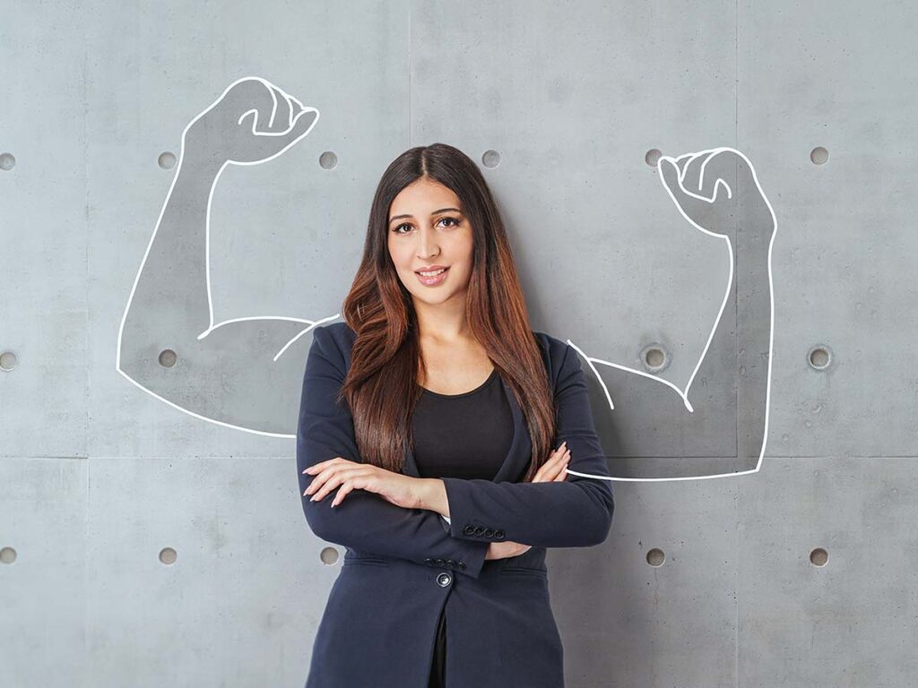Frau im Business-Outfit vor grauer Wand mit gezeichneten starken Muskeln, die Stärke und Selbstbewusstsein symbolisieren.