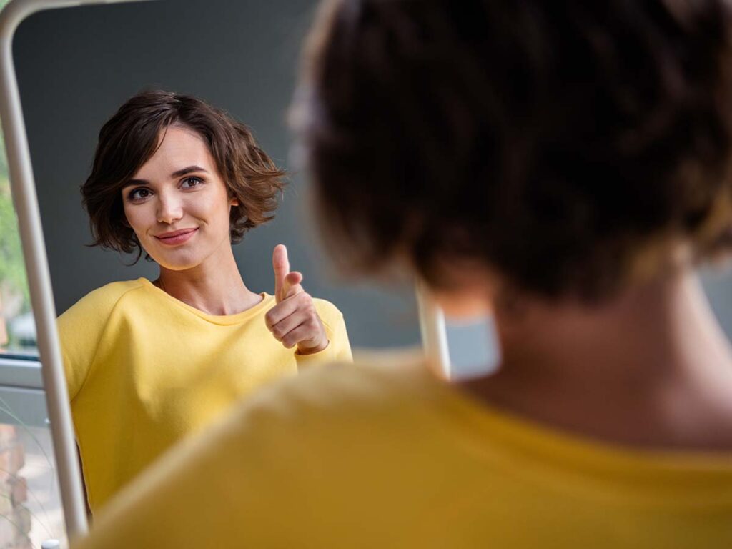 Frau im gelben Pullover blickt lächelnd in den Spiegel, zeigt auf sich und vermittelt Selbstbewusstsein und Motivation.