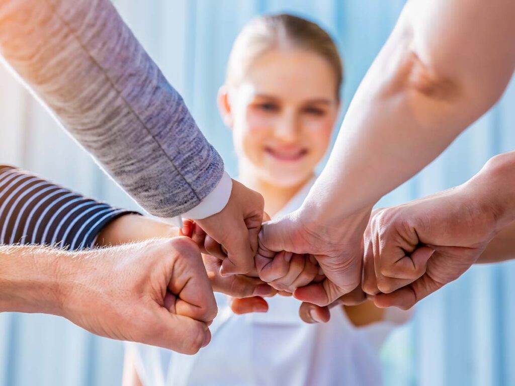 Verschiedene Hände im Kreis mit Fäusten zusammen, im Hintergrund ein lächelnder Mensch, symbolisieren Teamgeist und Zusammenarbeit.