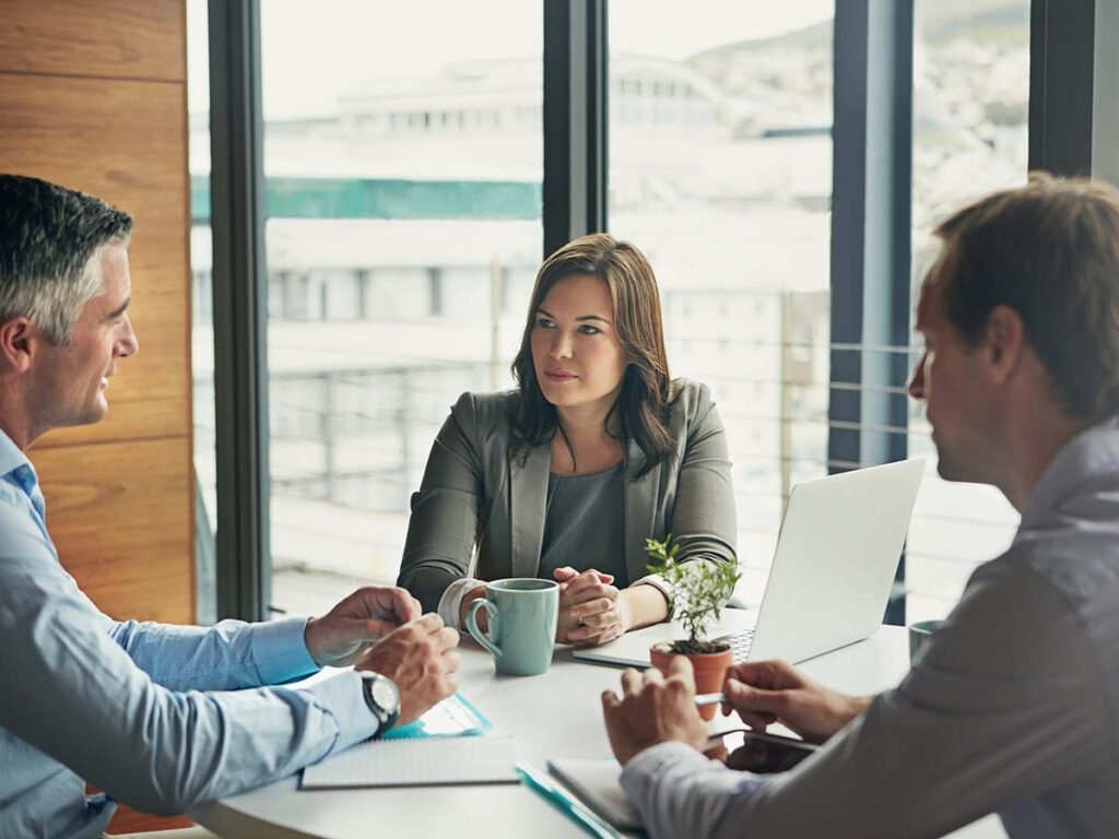 Besprechung im Büro: Drei Personen diskutieren am Tisch mit Laptop und Kaffeetasse, Fenster im Hintergrund, Geschäftstreffen.