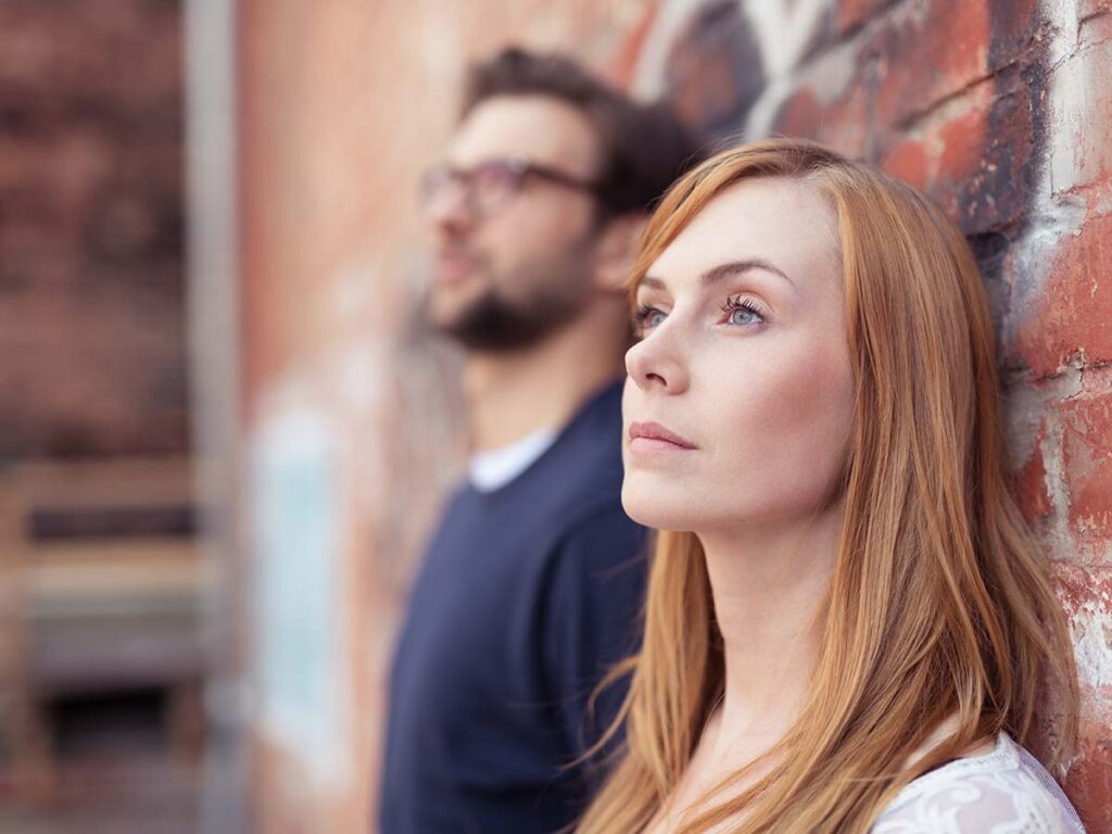 Frau mit roten Haaren lehnt nachdenklich an einer Backsteinmauer, unscharfer Mann im Hintergrund. Urbanes Porträtfoto.