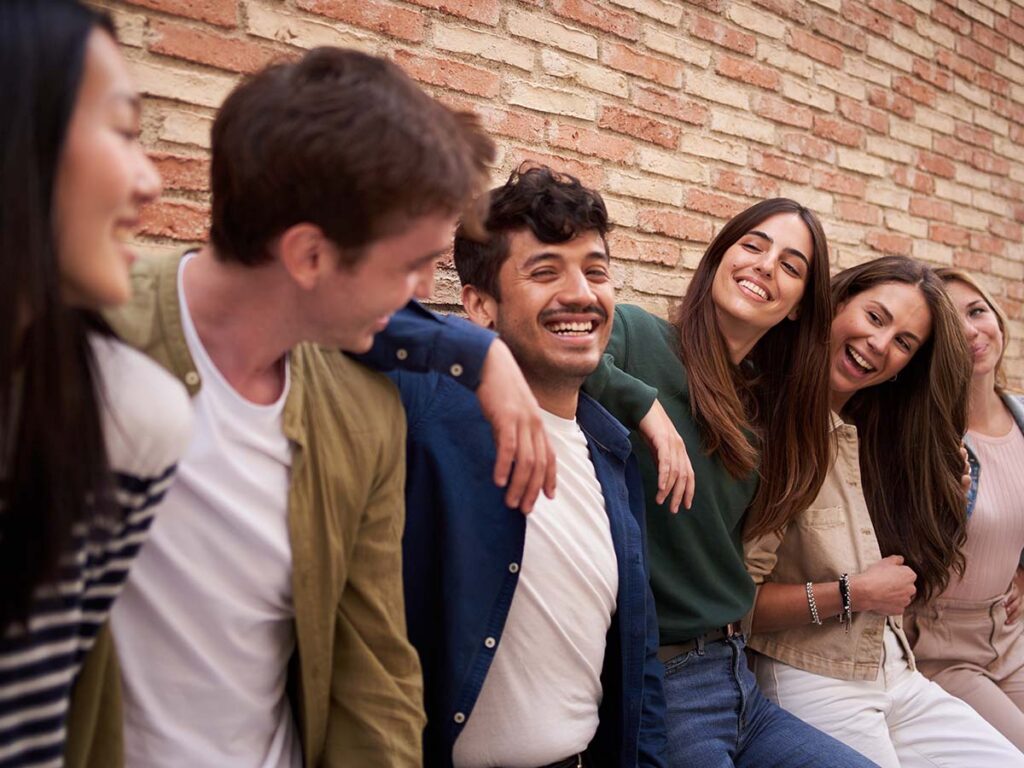 Gruppe fröhlicher Freunde lehnt an Backsteinmauer und lacht gemeinsam, symbolisiert Freundschaft und positive Stimmung.