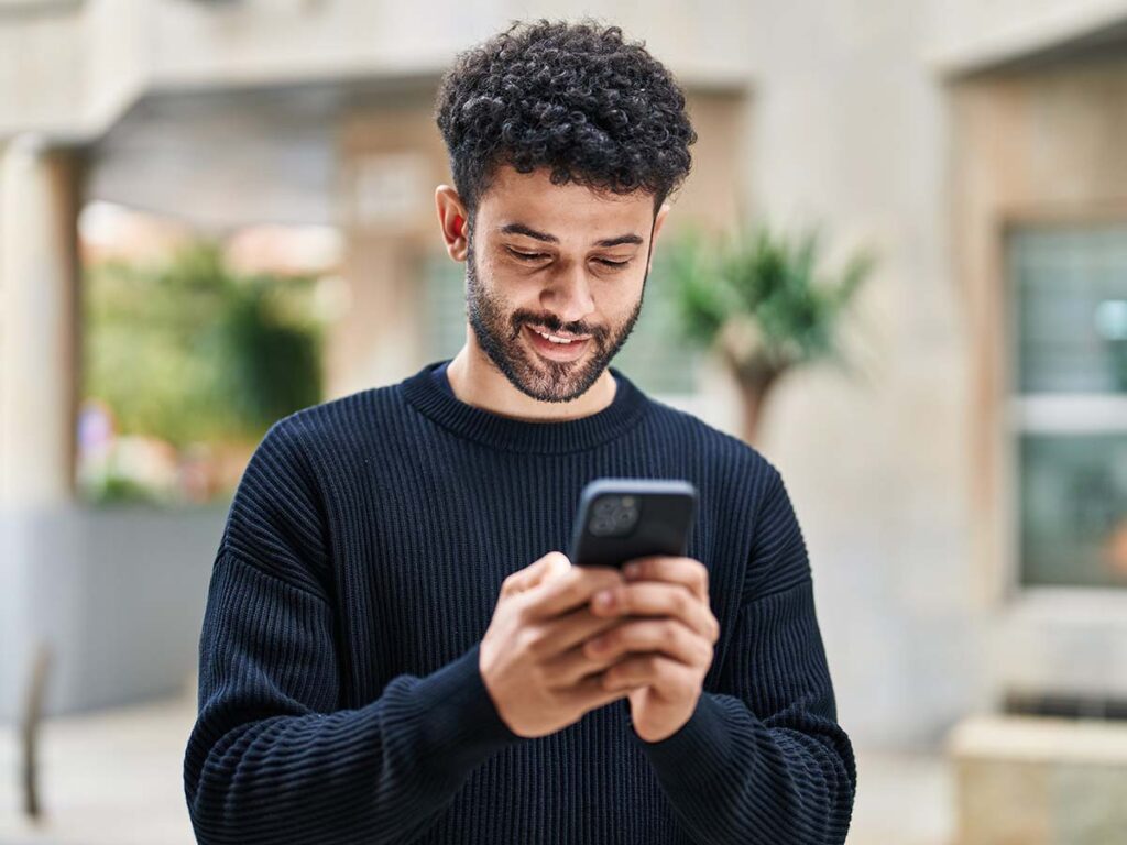 Junger Mann lächelt, während er auf Smartphone schaut. Freizeitkleidung, moderne Technologie, urbaner Hintergrund im Freien.