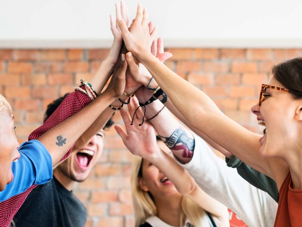 Gruppe von Freunden jubelt mit gemeinsamem High-Five vor einer Ziegelwand, symbolisiert Teamwork und Motivation.