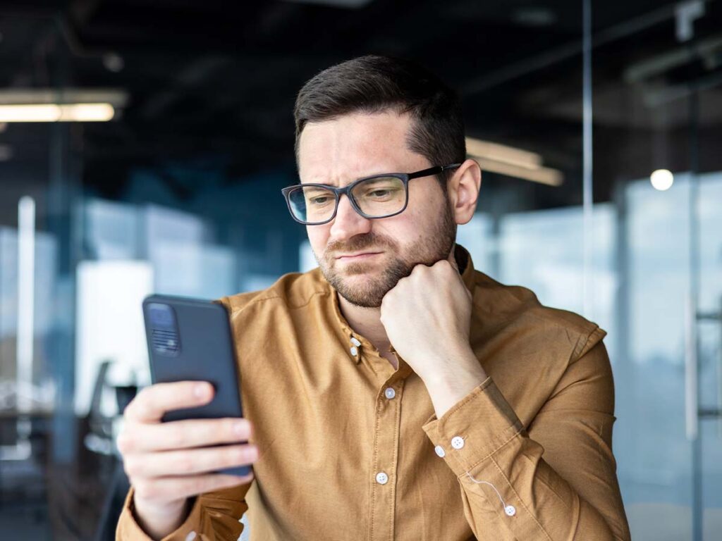 Mann mit Brille und Bart betrachtet nachdenklich sein Smartphone im Büro. Business-Kommunikation, Technologie, konzentriert.
