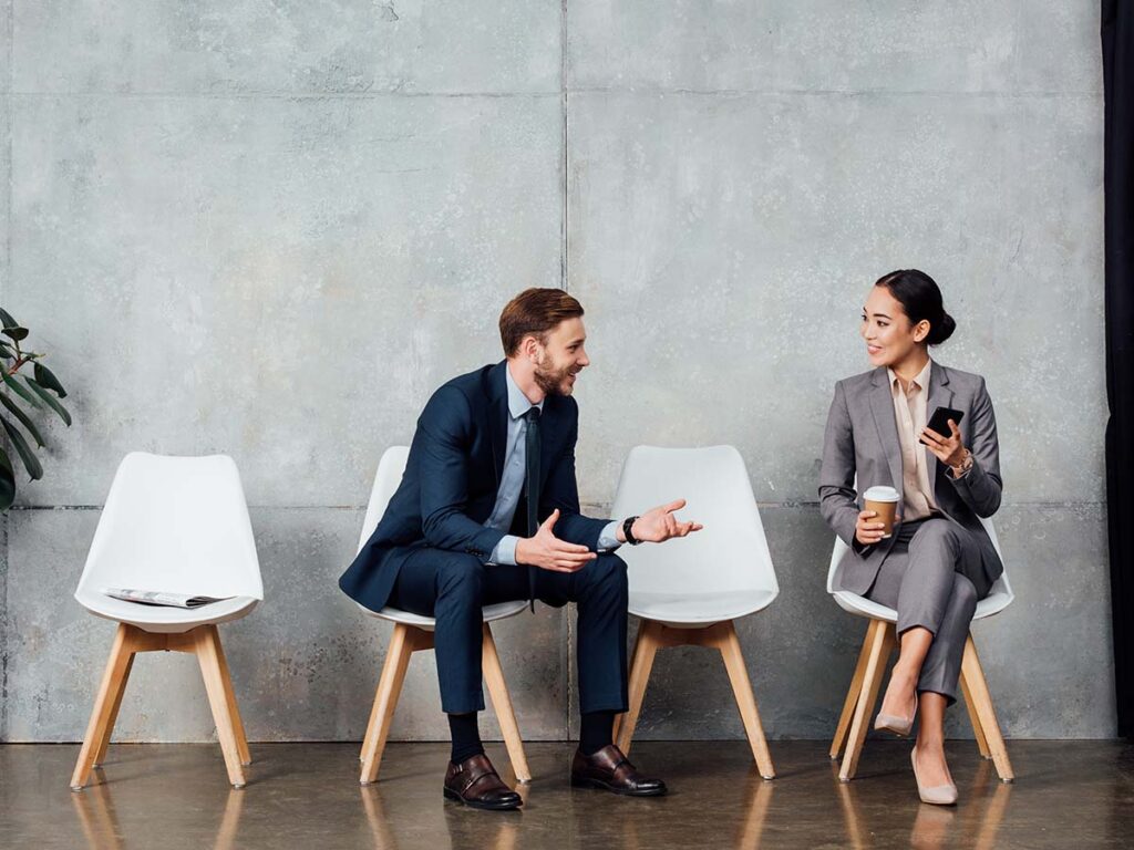 Zwei Geschäftsleute im Gespräch, sitzen auf Stühlen vor grauer Wand. Einer hält Handy und Kaffee, der andere gestikuliert.