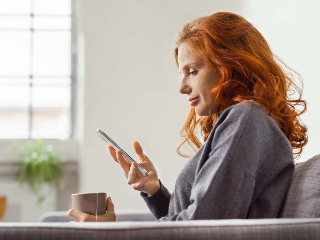 Frau mit roten Haaren trinkt Tee und benutzt Smartphone im Wohnzimmer. Gemütliche Entspannung zu Hause.