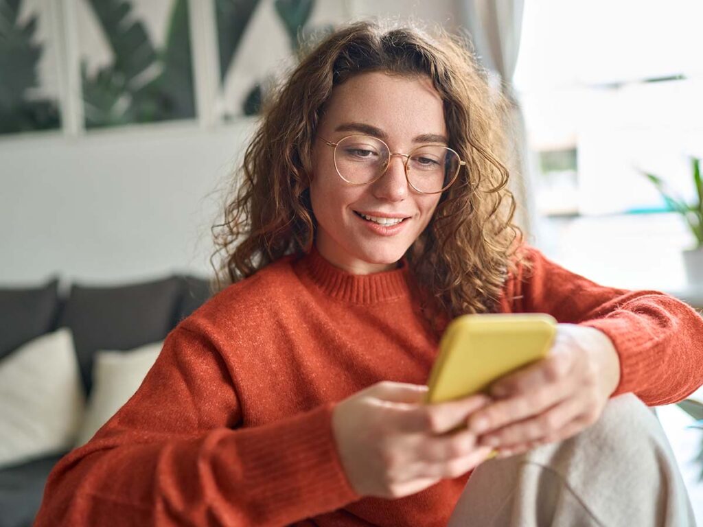 Junge Frau mit Brille und Locken lächelt beim Handybenutzen im Wohnzimmer. Entspannte Atmosphäre. Moderne Kommunikation.