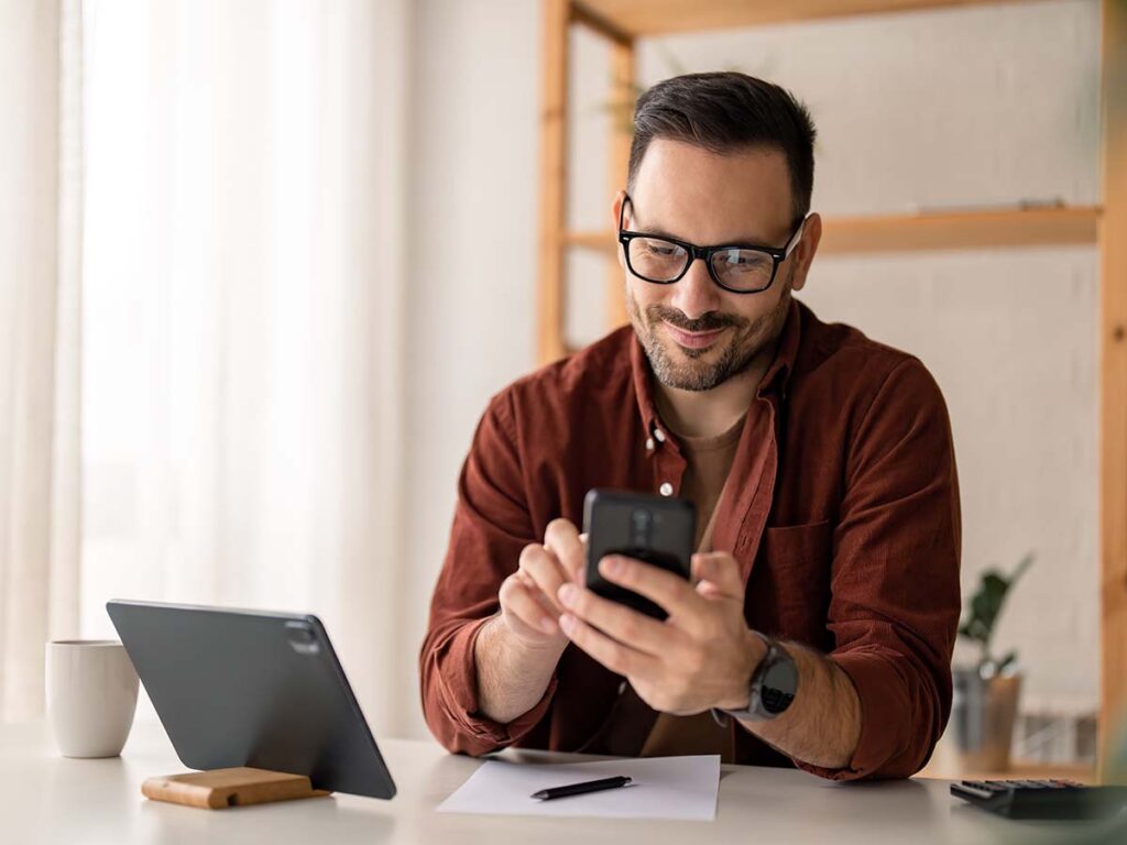 Mann mit Brille nutzt Smartphone am Schreibtisch mit Tablet. Modernes Arbeiten, Technologie, Homeoffice.