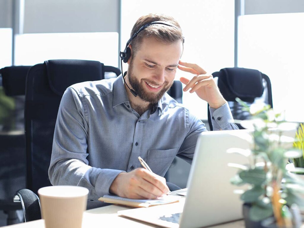 Mann im Büro arbeitet lächelnd am Laptop mit Headset, notiert auf Papier. Fokus auf Kommunikation, Kundenservice und Produktivität.