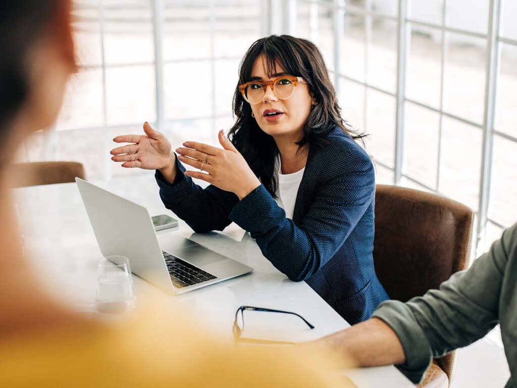 Besprechungsszene: Frau in Brille erklärt gestikulierend, vor Laptop im modernen Büro. Teamwork und Kommunikation im Fokus.