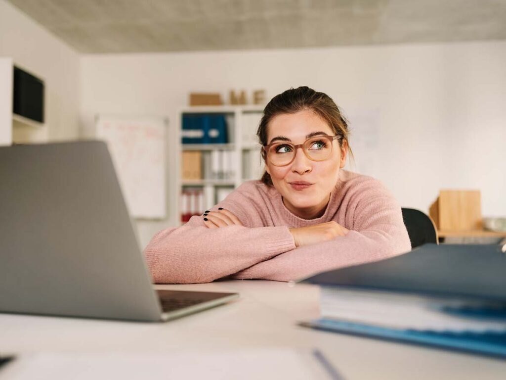 Eine Frau in rosa Pullover lächelt verträumt am Schreibtisch mit Laptop im Büro. Kreative Arbeitsumgebung im modernen Büro.