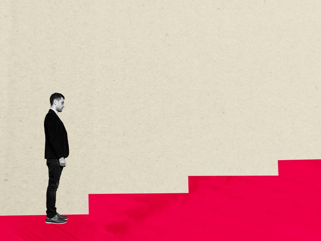 Mann steht vor roten Treppen auf grauem Hintergrund, symbolisiert Aufstieg. Herausforderung, Fortschritt, Karriere.