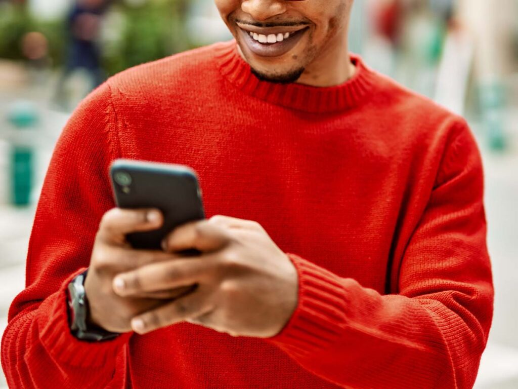 Mann in rotem Pullover lächelt beim Schreiben auf Smartphone im Freien. Technologie und Mobilität im Alltag.