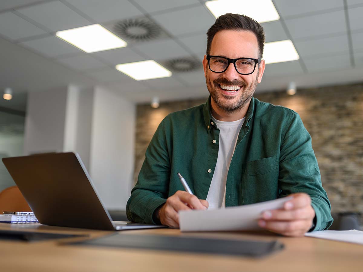 Lächelnder Mann in einem modernen Büro, der ein Dokument überprüft, symbolisiert die professionelle Zusammenarbeit im Dreiecksverhältnis der Zeitarbeit zwischen Zeitarbeitsunternehmen, Kundenunternehmen und Zeitarbeitnehmer.