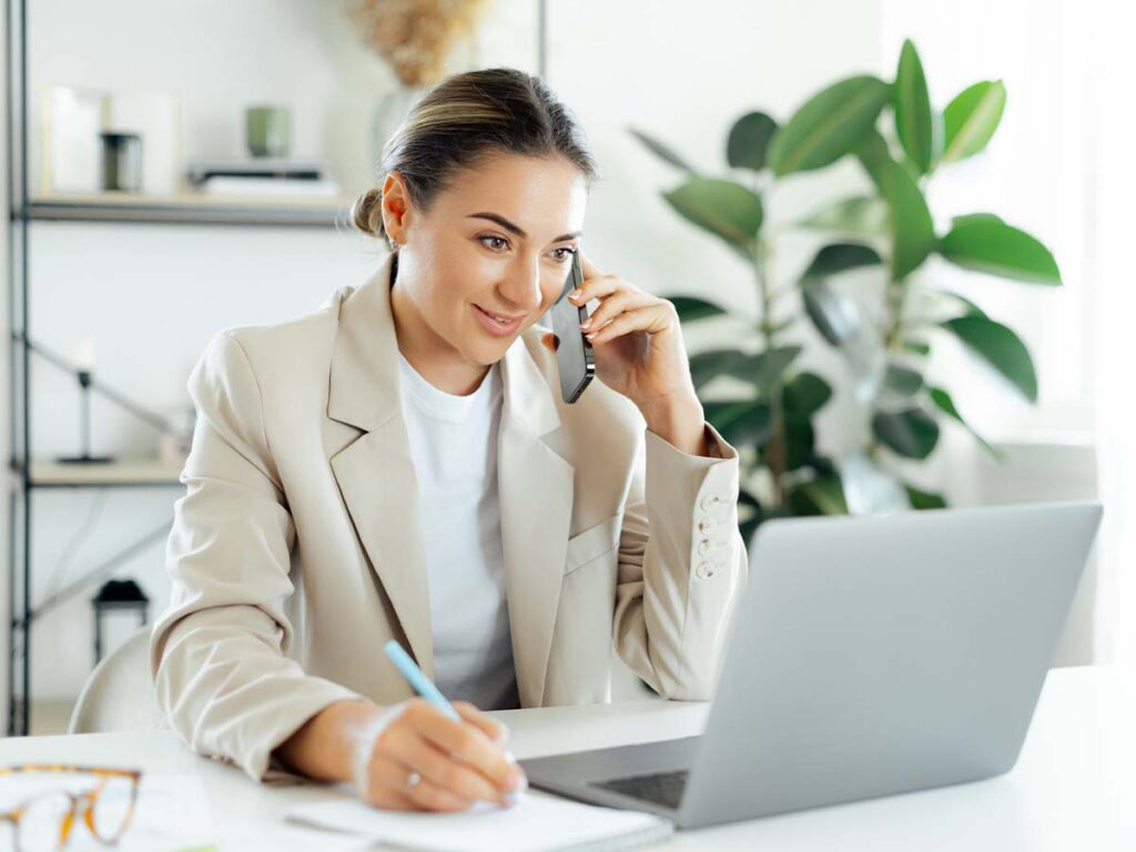 Eine Frau arbeitet am Laptop, führt ein Telefonat und notiert etwas. Homeoffice, Produktivität, effizientes Arbeiten.