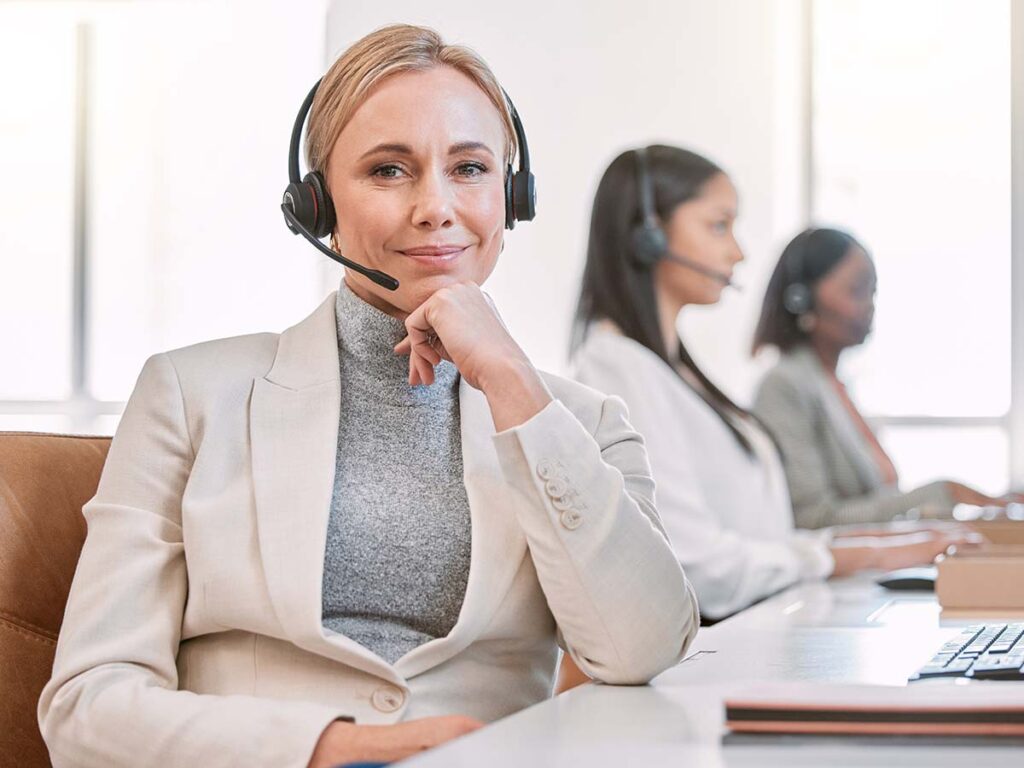 Lächelnde Frau mit Headset im Callcenter, Kundenservice, ehrgeizig. Zwei Kolleginnen arbeiten im Hintergrund.