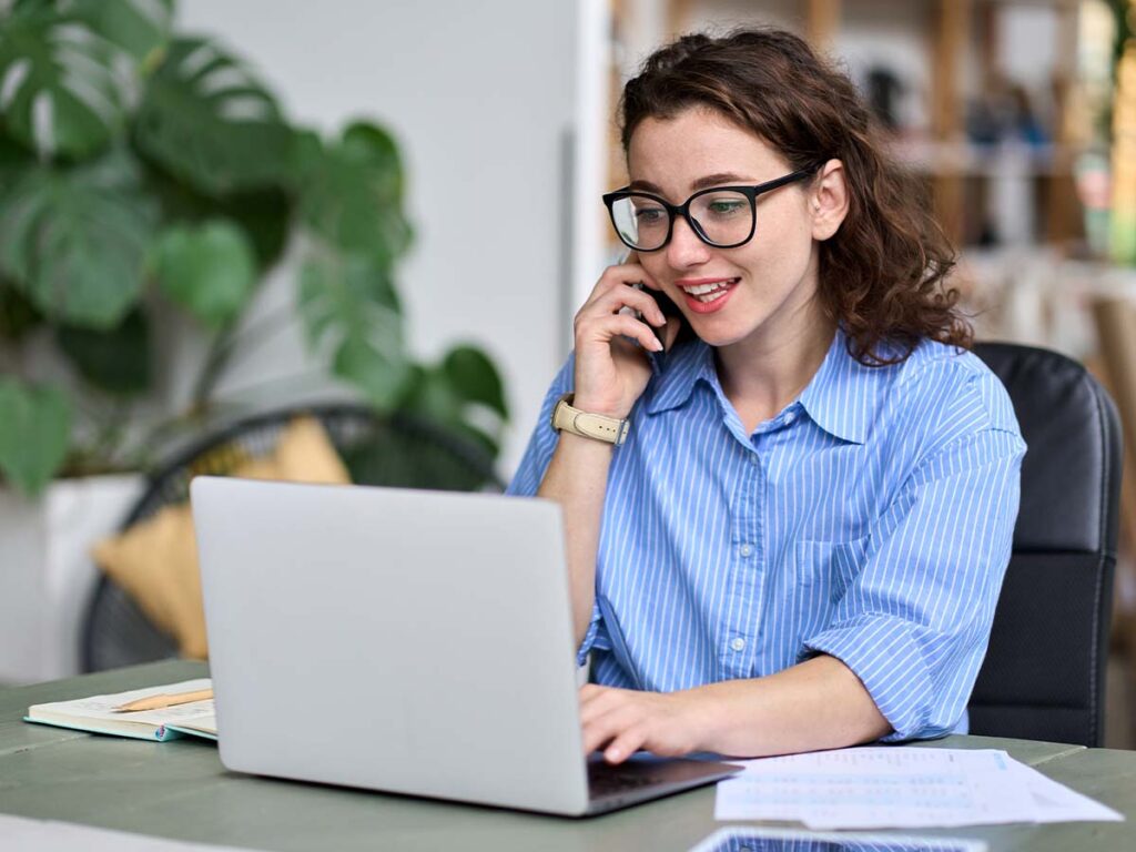junge Geschäftsfrau, professionelle Mitarbeiterin, oder Unternehmerin, die am Schreibtisch sitzt, online arbeitet auf dem Laptop, auf dem Handy, und spricht mit Kunden über Online-Website-Verkaufsauftrage oder Marketingarbeit.