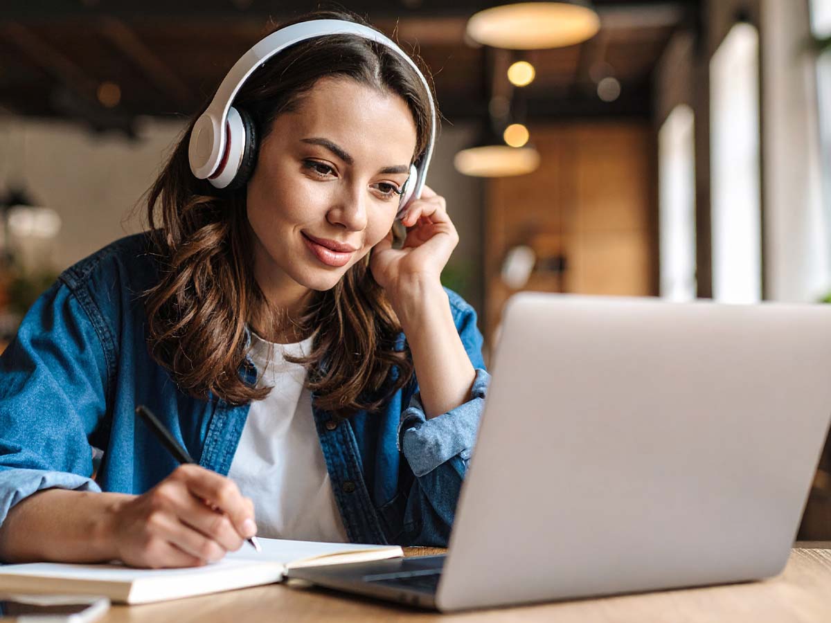 Junge Frau mit Kopfhörern lernt konzentriert online an einem Laptop und macht Notizen – modernes E-Learning in gemütlicher Umgebung