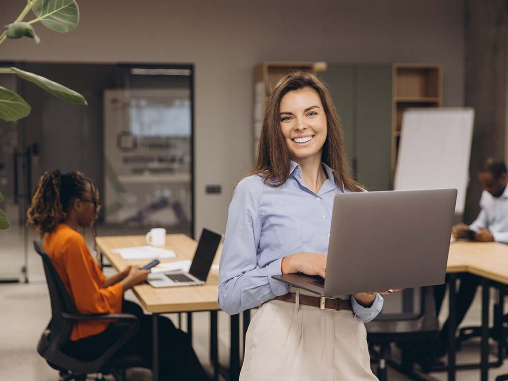 Büroangestellte mit Laptop lächelt, während Kollegen arbeiten. Moderne Arbeitsumgebung, Teamwork, Business-Konzept.