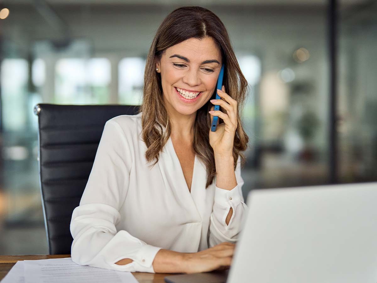 Eine Frau telefoniert im Büro und lächelt. Sie trägt eine weiße Bluse. Keywords: Kommunikation, Business, Laptop.